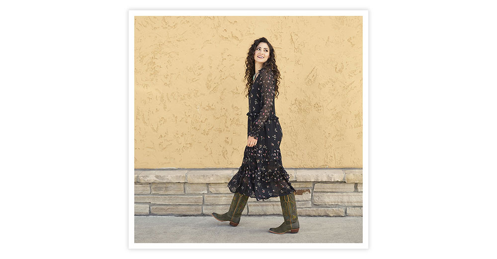 A woman with brown curly hair wearing a dress and Clara 15” western boots in green